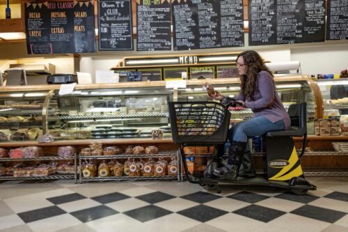 A woman in a mobility scooter examines a jar in front of a deli counter filled with pastries.