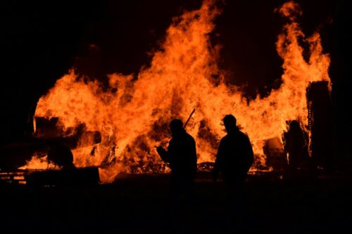 Silhouettes of two people stand before a large, intense wildfire at night. The flames are bright and towering, creating a dramatic scene.
