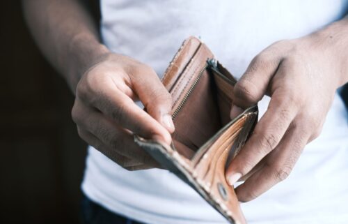 A person in a white shirt holds an open, empty brown wallet with both hands, conveying a sense of financial hardship, inflation, and job cuts.