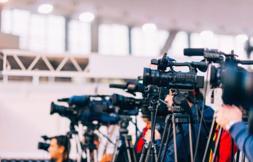 Rows of media with professional video cameras on tripods are lined up in a brightly lit indoor space. The atmosphere is focused, suggesting a press event.