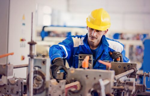 A worker in a blue uniform and yellow hard hat operates machinery in a factory. Focused expression, showcasing industrial labor and precision work.