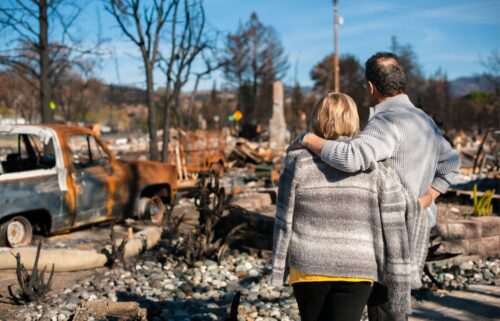 A couple in sweaters stands amidst a burned landscape, embracing and looking at the debris of a destroyed home and rusted truck, conveying loss and resilience.