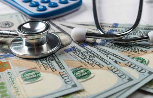 A stethoscope rests on several U.S. 100-dollar bills, with a calculator in the background, symbolizing healthcare costs and financial considerations.