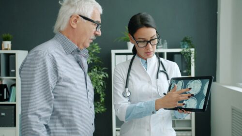 A female doctor in a white coat shows brain scans on a tablet to an older man in a checkered shirt. They are in a modern office, discussing intently. https://unsplash.com/photos/doctor-shows-medical-scan-to-elderly-patient-Pg4bY_O1kh8?utm_source=unsplash&utm_medium=referral&utm_content=creditShareLink