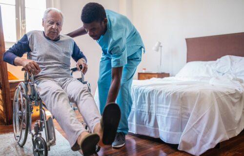 A caregiver in blue scrubs assists an elderly man in a wheelchair.