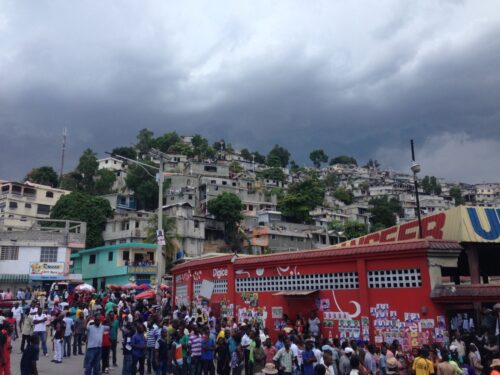 Crowded street scene in Haiti as voters gather outside a polling center during the country's August 2015 elections. Dense hillside housing and dark storm clouds loom overhead, creating a tense atmosphere.