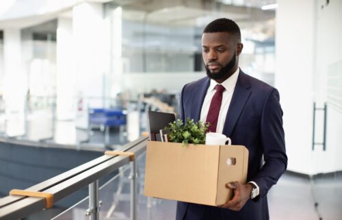 A man in a suit carries a box with office items like a plant and mug, walking through a modern office with a serious expression, suggesting job loss.