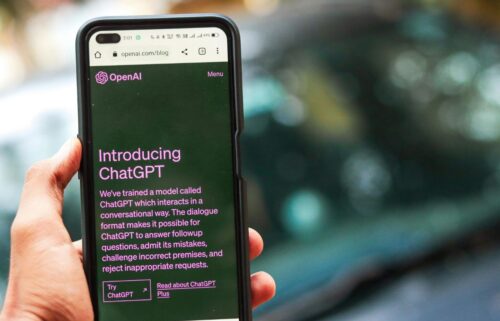 A hand holds a smartphone displaying the ChatGPT website. The screen shows text introducing ChatGPT by OpenAI with green and purple tones.