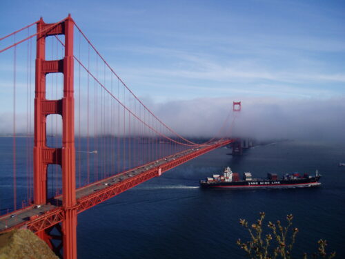 The Golden Gate Bridge in California stretches over calm blue waters. A container ship sails beneath, and thick fog partially obscures the background, creating a serene, majestic atmosphere.