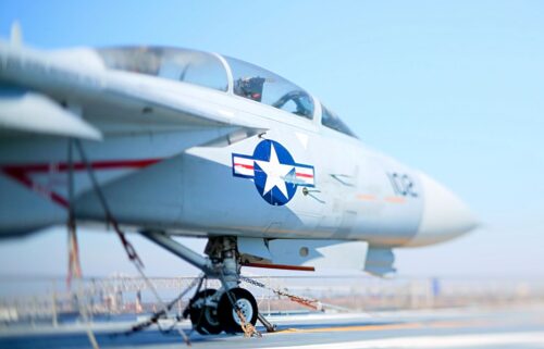 A military jet with a US Air Force emblem rests on a runway. The aircraft is secured with chains. The sky is clear, creating a calm atmosphere.