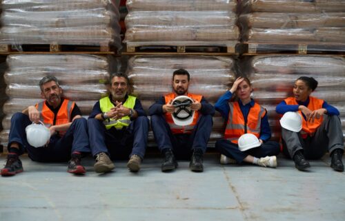 Five union workers in orange safety vests and helmets sit on the floor of a warehouse, leaning against stacked pallets, appearing tired yet relaxed.