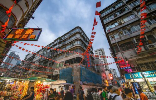 Bustling China street scene with vibrant red banners strung between tall, weathered buildings. Neon signs glow, and a busy crowd fills the marketplace below.