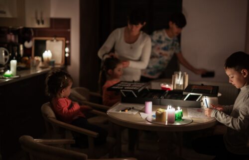 A family in dim lighting during a power outage, lit by candles on a table. Children draw while an adult assists.