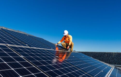 A worker, wearing an orange safety vest and helmet, crouches on a roof, installing solar panels under a clear blue sky. The scene conveys innovation and sustainability.