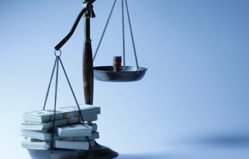 A balance scale is weighed heavily on one side with stacks of paper money, while the other side holds a few coins, set against a blue background representing inequality.