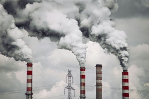 Four tall chimneys spewing thick, white smoke into a cloudy sky, conveying industrial pollution. A metal power line tower stands nearby. https://unsplash.com/photos/smoke-billows-from-the-stacks-of-smoke-stacks-SvOolOyZQPc?utm_source=unsplash&utm_medium=referral&utm_content=creditShareLink