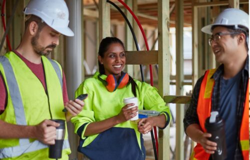 Three construction workers in safety gear, including helmets and vests, share a cheerful conversation at a building site. They hold coffee cups and lunch.