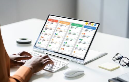 A person types on a laptop displaying a digital Kanban board with columns: To Do, In Progress, Testing, Done. The scene suggests productivity.