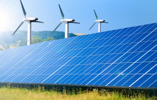 A field of blue solar panels is in the foreground with three wind turbines in the background against a clear blue sky, symbolizing renewable energy.