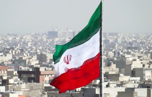 A large Iranian flag waves prominently against a hazy skyline of densely packed urban buildings, symbolizing national pride and urban life.