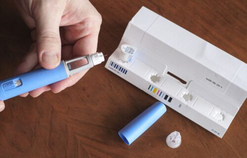 A hand holds a blue Ozempic pen beside its packaging on a wooden table. The pen cap and needle tips are visible, suggesting preparation for use.