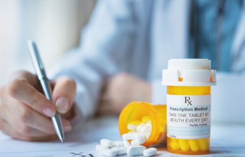 Close-up of a doctor's hand holding a pen near prescription paperwork. A pill bottle labeled 