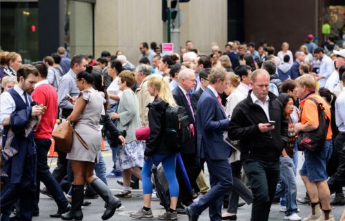 A bustling city street with a diverse crowd crossing. People wear business and casual attire, some on phones, conveying a busy, energetic urban atmosphere.