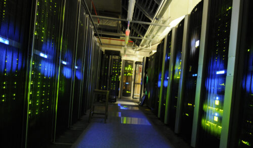 Dimly lit AI data center corridor with rows of server racks on both sides, glowing with green and blue LED lights, creating a futuristic ambiance.