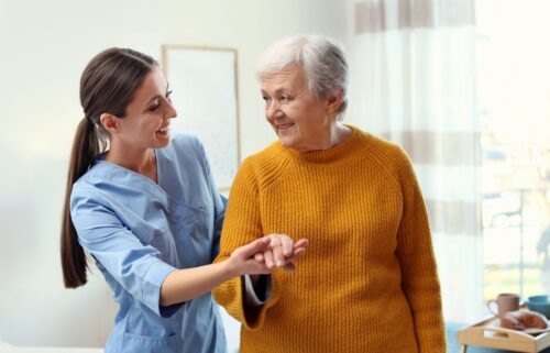 A medicare nurse in blue scrubs smiles while supporting an elderly woman in a mustard sweater. They hold hands warmly in a sunlit room, conveying care and joy.