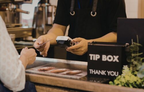 A barista in a black apron processes a card payment at a coffee shop counter. A 