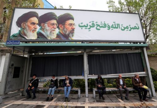 Iranian sit at bus stop beneath a billboard depicting Iran's late supreme leader Ayatollah Ruhollah Khomeini (L), his successor the late Ayatollah Ali Khamenei (C) and the new supreme leader Mojtaba Khamenei in Tehran on March 31, 2026. US and Israeli strikes hit military facilities in central Iran, damaged a major religious site in the northwest and provoked power cuts on March 31, after the US president threatened to blow up the country's oil and energy plants. (Photo by ATTA KENARE / AFP via Getty Images) /