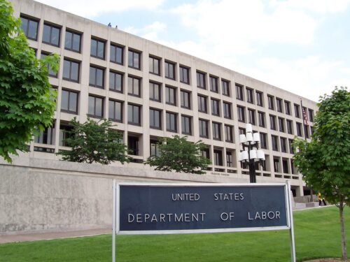 A large, rectangular building with rows of windows, surrounded by green trees. In front, a sign reads 