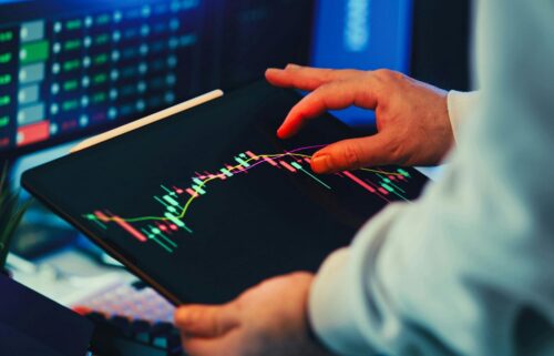 Hands holding a tablet displaying a financial stock chart with colorful candlesticks. Monitors in the background suggest a trading environment.
