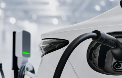 Close-up of an electric vehicle being charged, with a black charging cable attached. Background features a softly blurred, bright indoor setting.