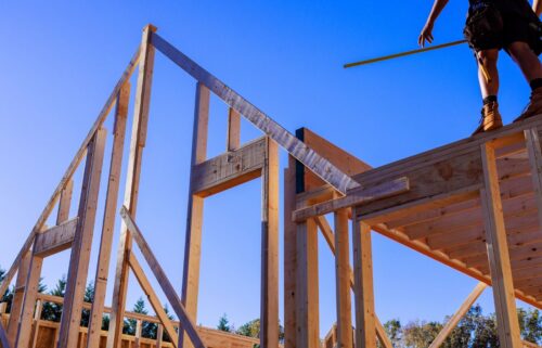 Wooden house frame under construction against a clear blue sky; a person stands on top holding a yellow measuring tape, conveying progress.