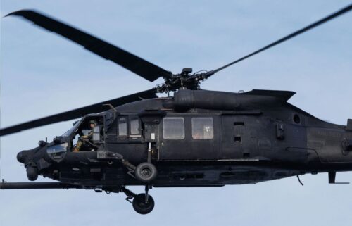 A black military helicopter, mid-flight against a clear blue sky, with a person visible in the cockpit. The rotor blades are spinning dynamically.