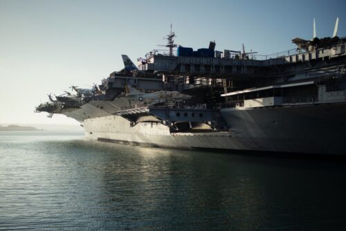 Military aircraft carrier with planes ready on deck. The sun sets behind, casting reflections on the water. https://unsplash.com/photos/gray-warship-on-body-of-water-BQgAYwERXhs?utm_source=unsplash&utm_medium=referral&utm_content=creditShareLink