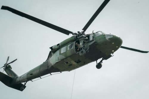 A military helicopter in flight against a cloudy sky, with a soldier visible inside. The helicopter's rotors are captured in motion, conveying urgency.