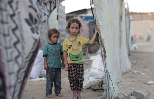 Two young children stand in a makeshift shelter with dirt ground. The girl wears a yellow shirt; the boy is in blue. Their expressions are solemn.