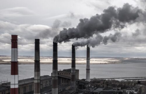 Industrial smokestacks emit thick gray smoke against a cloudy sky, overlooking a body of water. The scene conveys pollution and environmental impact.