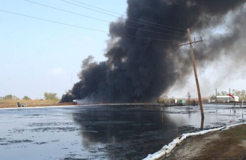 Thick black smoke billows from a fire near a flooded field. A leaning telephone pole is in the foreground, conveying urgency and chaos.
