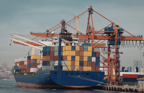 A large cargo ship at a busy port, loaded with colorful stacked containers. Red cranes surrounding it. City skyline in the hazy background.