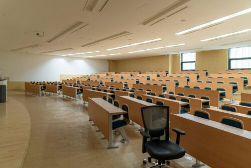 Spacious, empty lecture hall with rows of wooden desks and blue chairs. A single black chair stands in the foreground.