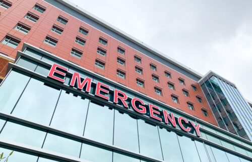 View of a hospital's emergency department. Large red letters spelling “EMERGENCY” are on the building's glass front, conveying urgency against a cloudy sky.