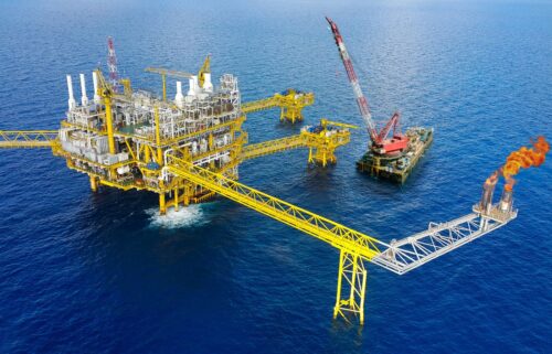 Aerial view of an offshore oil rig with yellow structures, surrounded by blue ocean. Flames emerge from a flare stack, and a crane is nearby.