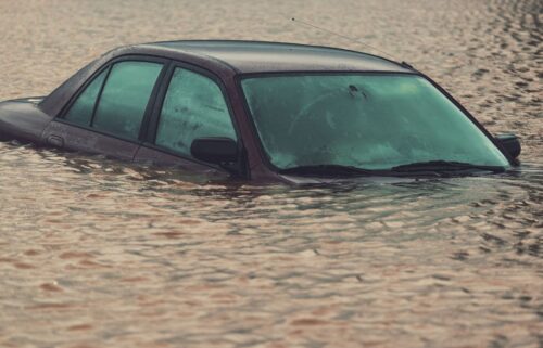 A partially submerged car in murky floodwaters, only its roof and windows visible. The scene conveys a sense of abandonment and disaster.