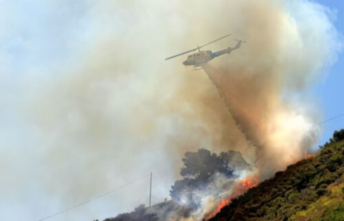 Helicopter drops water over a smoky wildfire on a hill, surrounded by thick smoke and flames, against a clear blue sky, conveying urgency and tension.