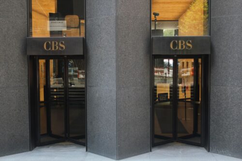 Two revolving doors of a CBS building entrance, flanked by gray stone walls. The glass reflects a city scene, giving a sleek and professional tone.