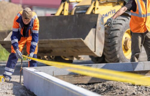 Two construction workers in safety gear measure with a tape along a metal beam at a work site, with heavy machinery in the background.