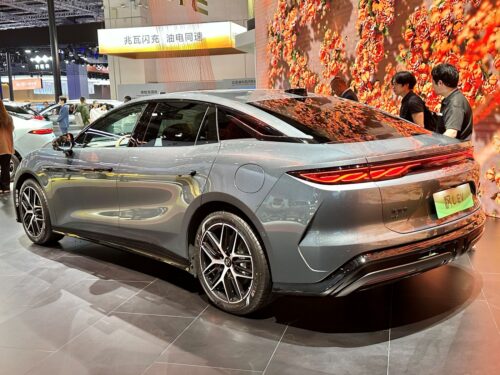 Sleek gray BYD electric car showcased indoors with vivid floral backdrop. People admire the vehicle's modern design and striking rear lights. Photo by JustAnotherCarDesigner https://commons.wikimedia.org/w/index.php?curid=164175644